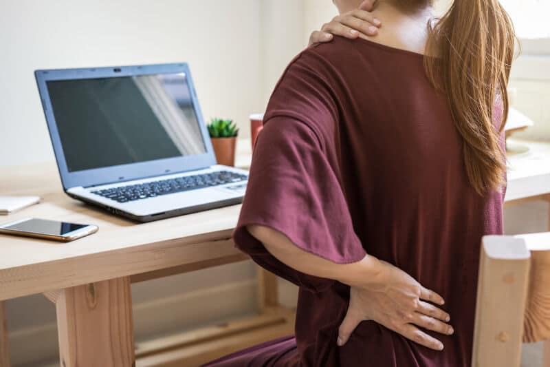 A woman sitting at a desk with a laptop holding her neck and lower back, indicating pain.